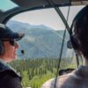 pilot and passenger look out the front window of a helicopter over green mountain landscape