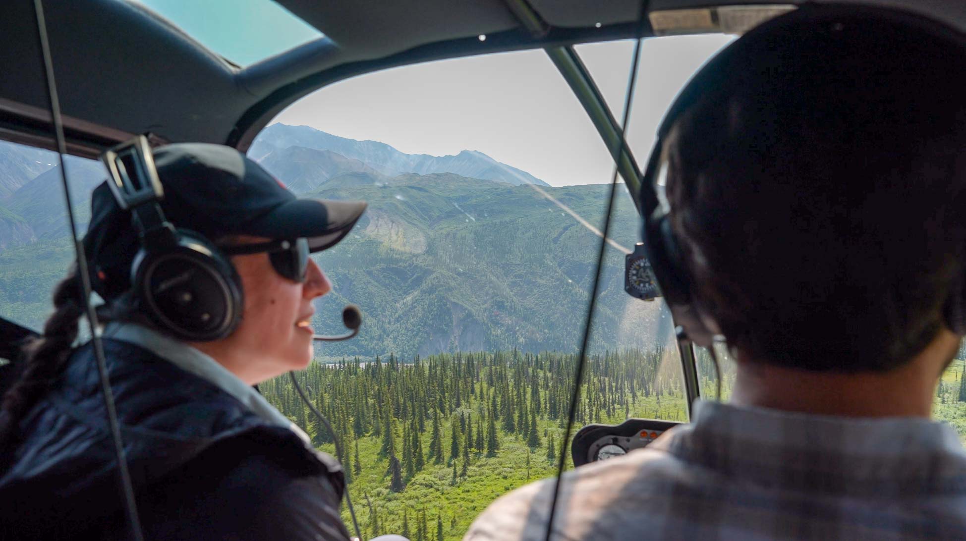 pilot and passenger look out the front window of a helicopter over green mountain landscape