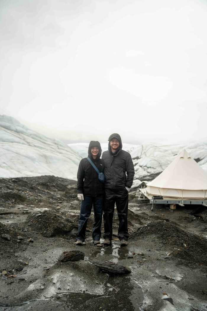 two people in front of canvas tent on glacier moraine with white ice and fog in background